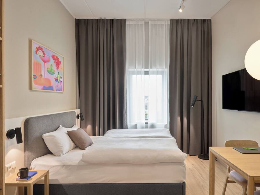 Quarto de hotel com cama, mesa de cabeceira, escrivaninha, cadeira, televisão e janela com cortinas.