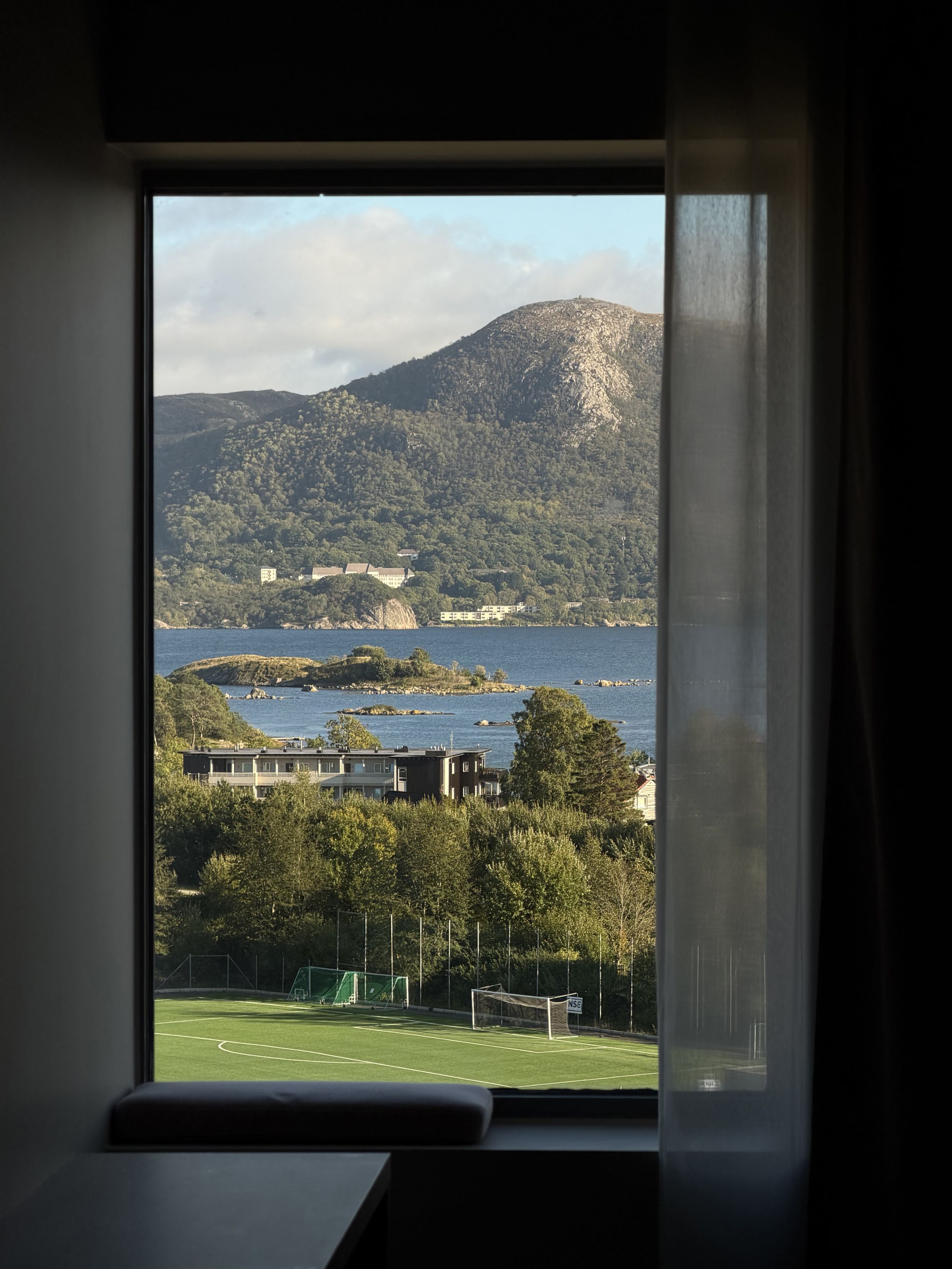 A window view of a coastal landscape with a mountain, a body of water, and a sports field.