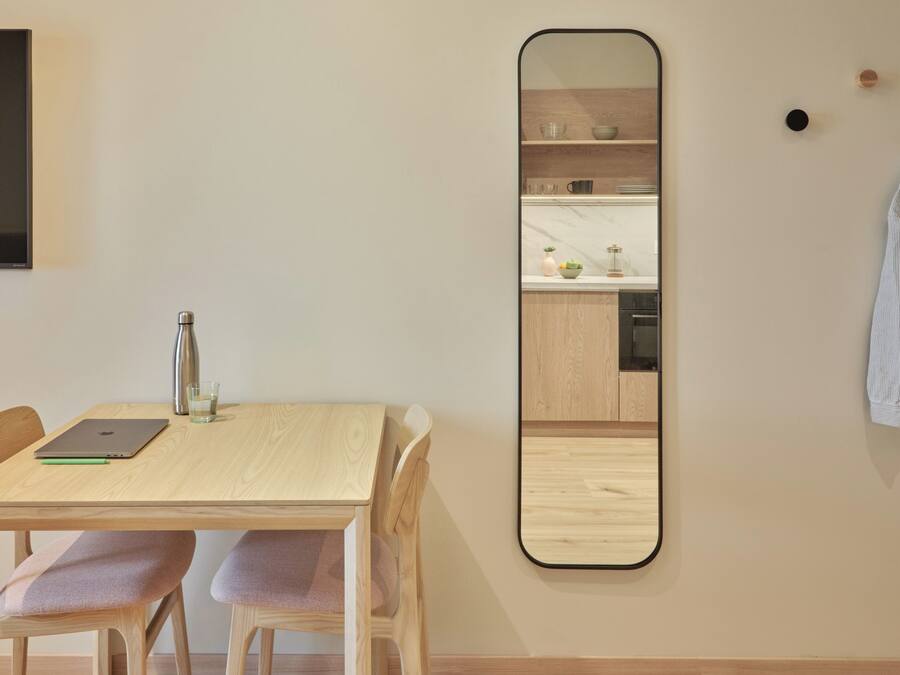 Un comedor con una mesa de madera, dos sillas, una computadora portátil, una botella de agua y un vaso. Un espejo grande está montado en la pared, reflejando la habitación.