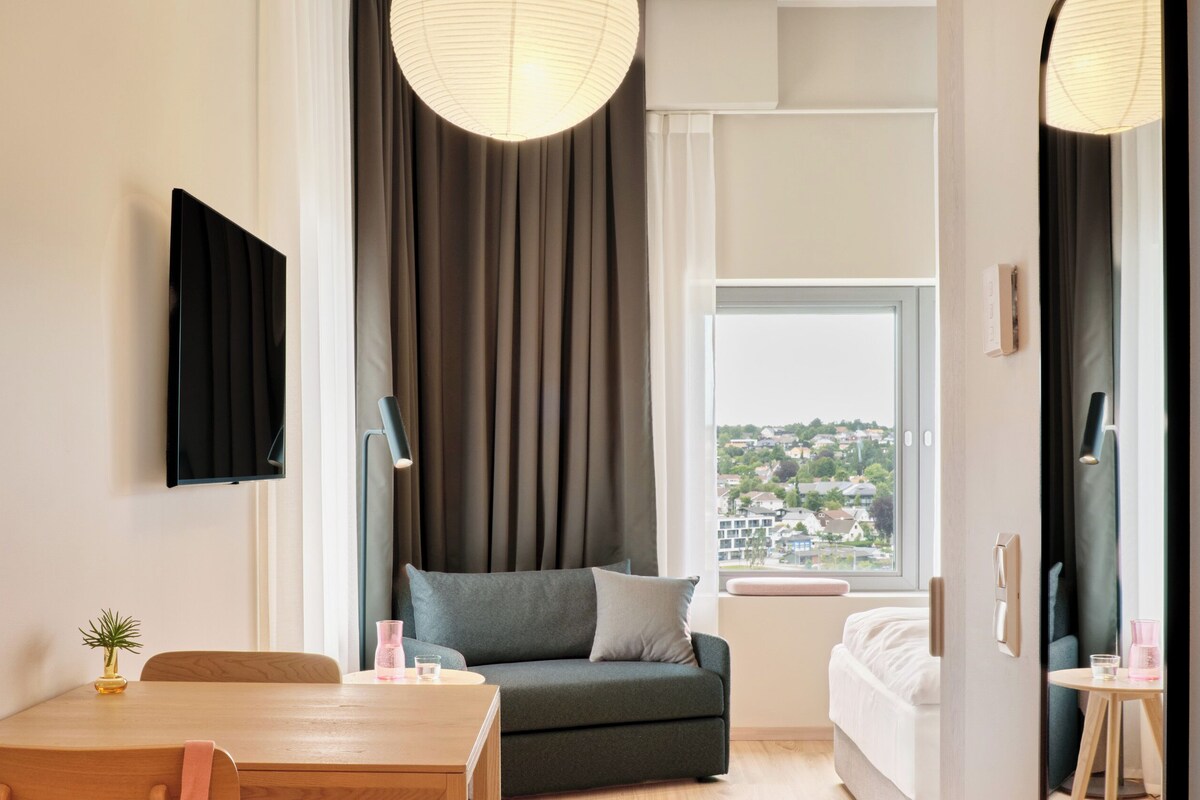 A modern hotel room with a dining table, sofa, and a view of a cityscape.