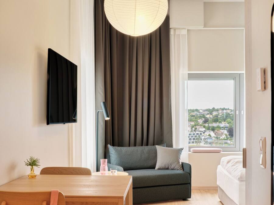A modern hotel room with a dining table, sofa, and a view of a cityscape.