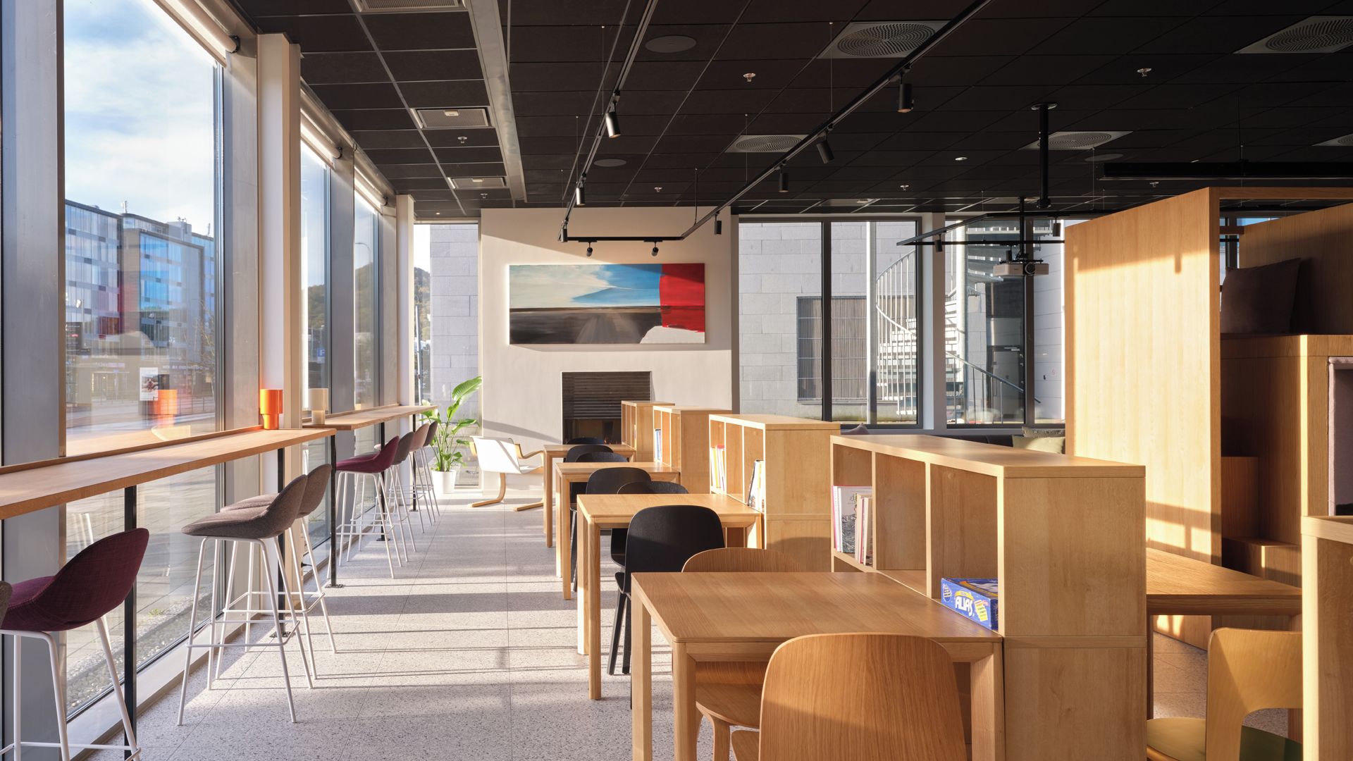 Una biblioteca moderna con mesas y sillas de madera, amplias ventanas y vistas al paisaje urbano.