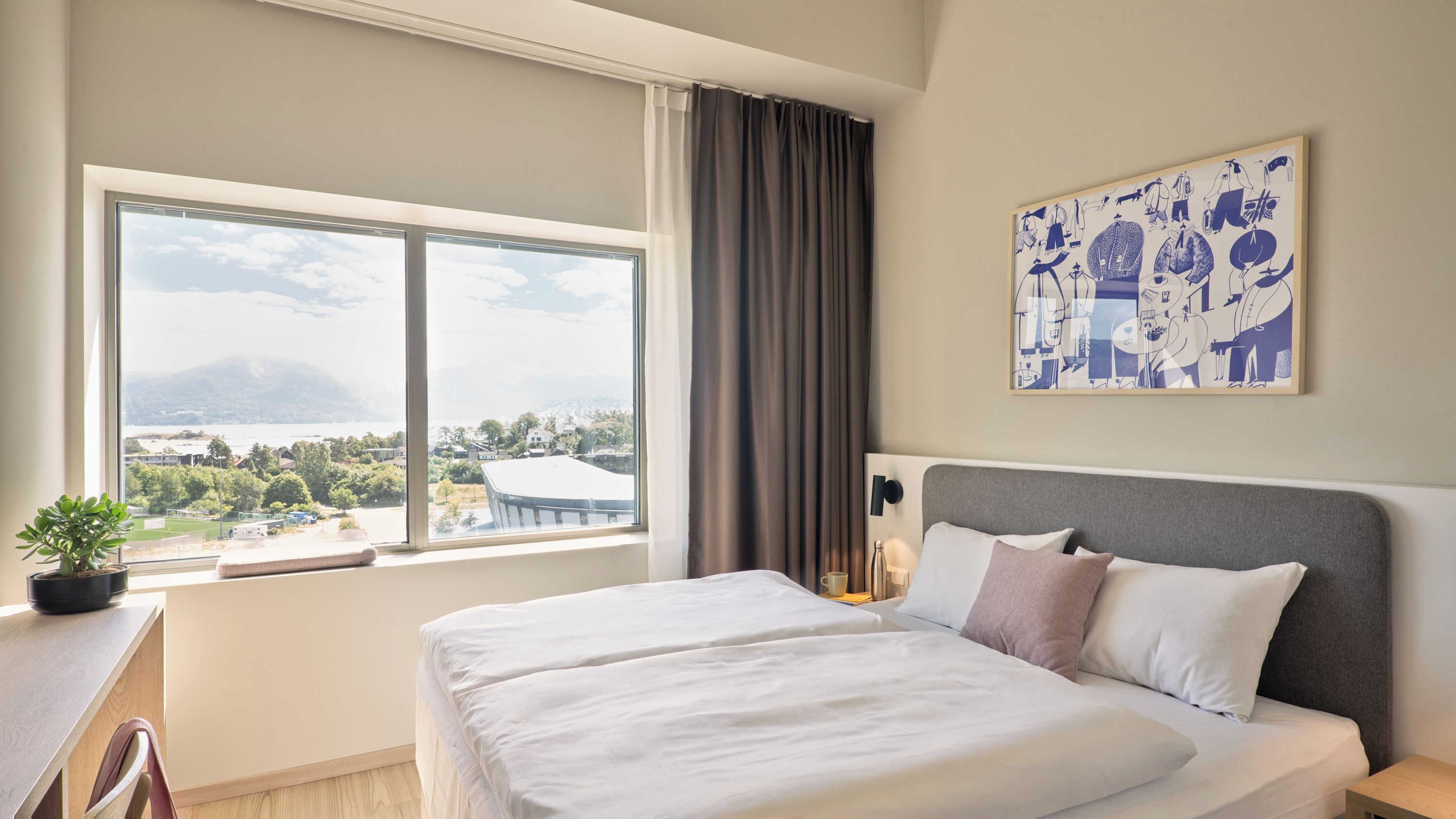 A hotel room with a large bed, a desk, a potted plant, a view of the outdoors, and a piece of wall art.