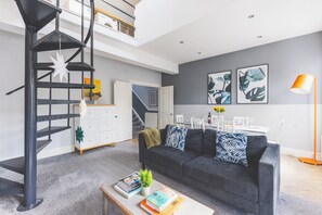 A modern living room with a staircase, a grey sofa, a coffee table with books, and a dining area in the background. - Designer Battersea Family Home | 3BR | Sleeps 6 (Greater London)