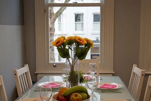 A dining table with a vase of flowers, a fruit bowl, and a window with a view of buildings.