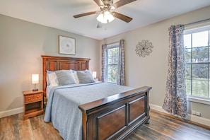 Une chambre à coucher avec un lit en bois, une table de chevet, un ventilateur de plafond et une fenêtre ornée de rideaux à motifs.