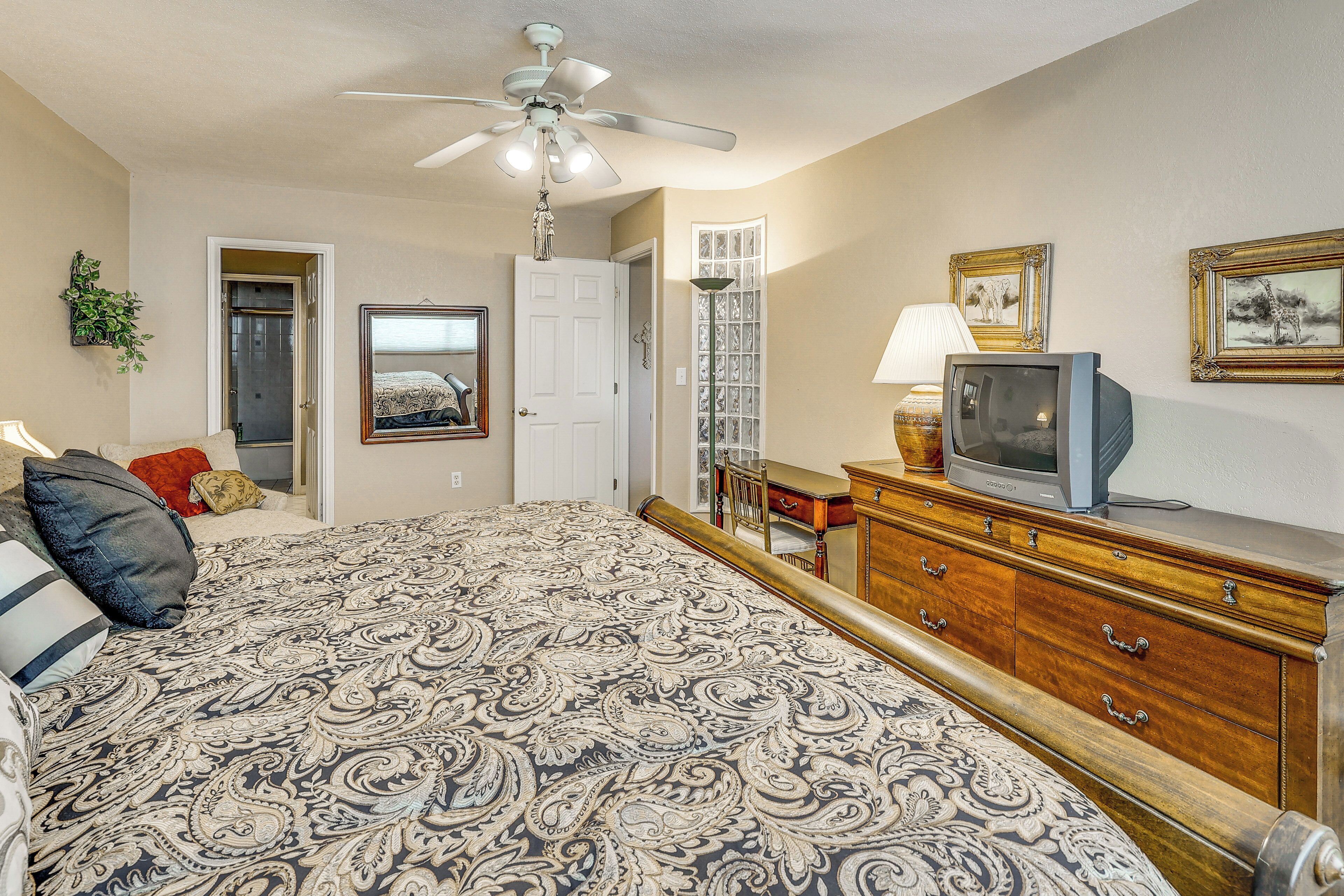 A bedroom with a patterned bedspread, a ceiling fan, a television on a dresser, and a door leading to another room.