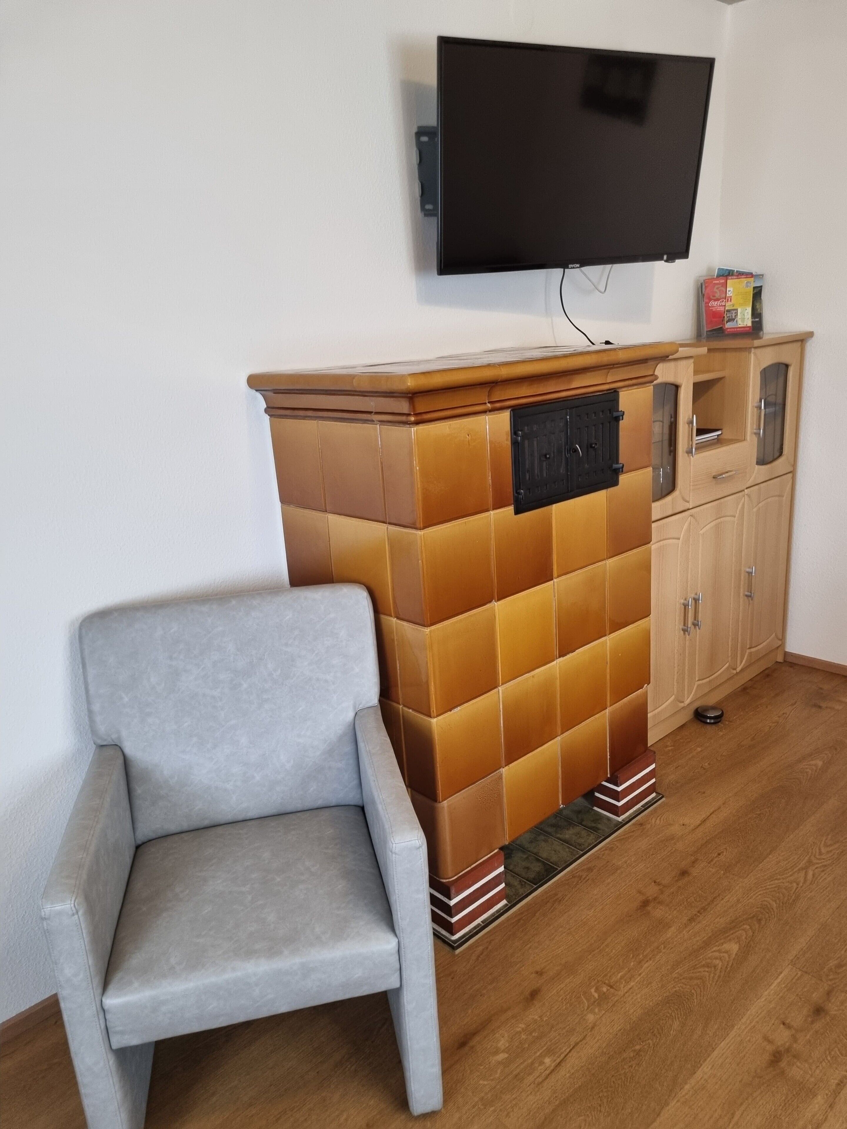 Une pièce comprenant un fauteuil gris, une cheminée recouverte de carreaux, un meuble en bois et un téléviseur à écran plat fixé au mur.