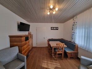 Ein Zimmer mit Holzboden, einem an der Wand befestigten Flachbildfernseher, einem Esstisch mit Stühlen, einer Couch und einem Fenster mit durchsichtigen Vorhängen.