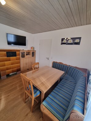 Ein Zimmer mit einem Holzesstisch, einer Couch, einem an der Wand montierten Fernseher und einer Vitrine.
