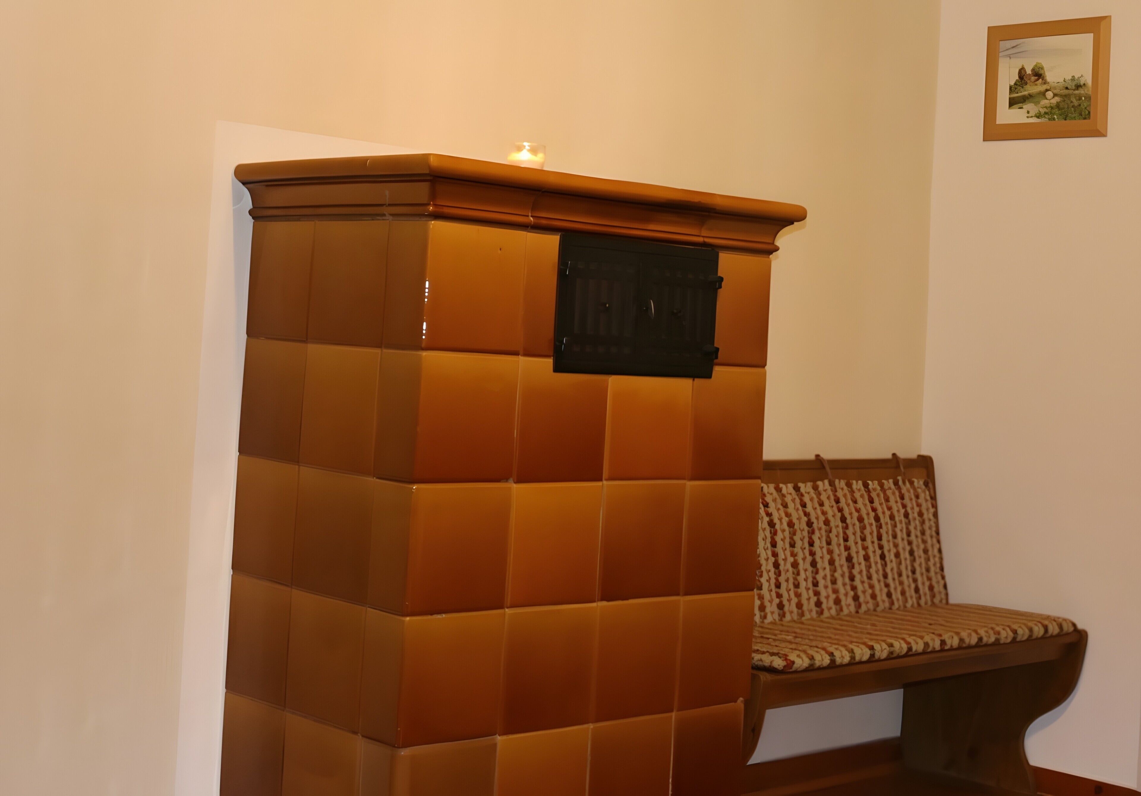 Een hoekje met een bruine, beklede bank, een tafeltje met een kaars en een lijst met een afbeelding aan de muur.