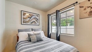 Ein Schlafzimmer mit einem Bett, einem Fenster mit Vorhängen, einem gerahmten Kunstwerk an der Wand und einer Tür.