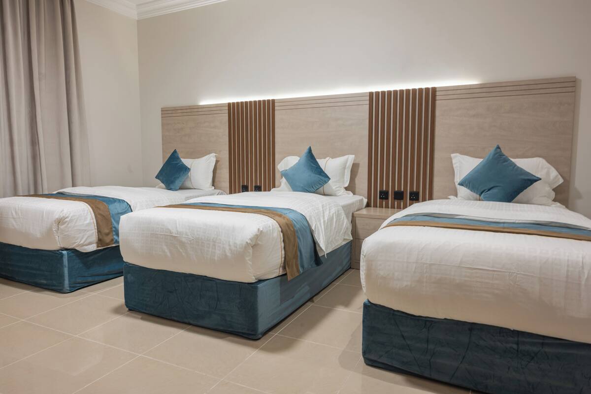 Une chambre d’hôtel avec trois lits, chacun doté d’une tête de lit bleue, de draps blancs et d’un élément décoratif marron.