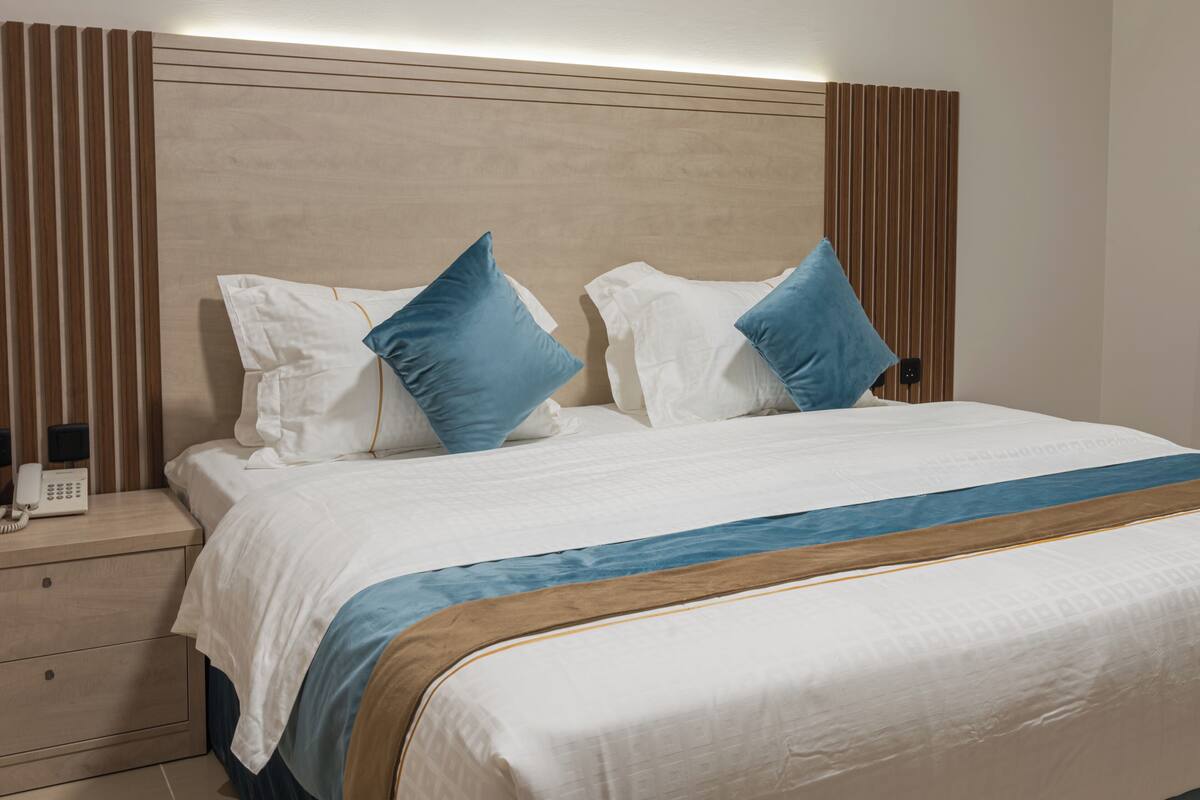 Un lit bien fait, avec une tête de lit en bois, une table de chevet sur laquelle se trouve un téléphone et un coussin bleu décoratif.