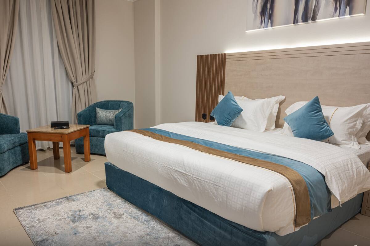 Une chambre moderne avec un grand lit, deux fauteuils, une petite table en bois et une grande fenêtre avec des rideaux.