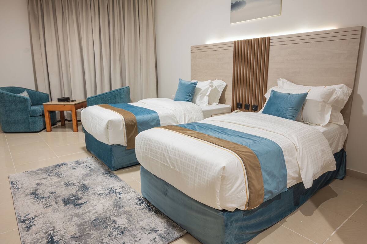 Une chambre d’hôtel avec deux lits, un fauteuil bleu, une petite table et une grande fenêtre avec des rideaux.