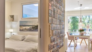 A bedroom with a bed, a nightstand, a lamp, a wall-mounted shelf, and a window with a painting. - LichtSpiel Scharbeutz - Arrive, take a deep breath, enjoy - Apartment LichtSpiel (Scharbeutz)