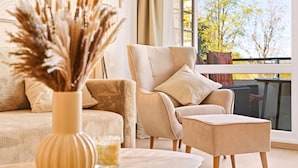 Ett modernt vardagsrum med en beige soffa, en tillhörande fåtölj och en liten pall. I förgrunden står en vas med torkade växter.