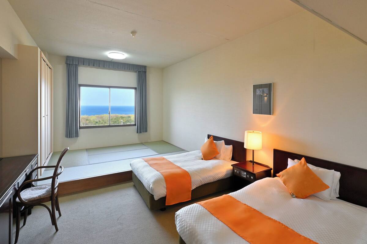 Une chambre d’hôtel avec deux lits, un bureau, une chaise, une fenêtre donnant sur un beau paysage et une table de chevet avec une lampe.