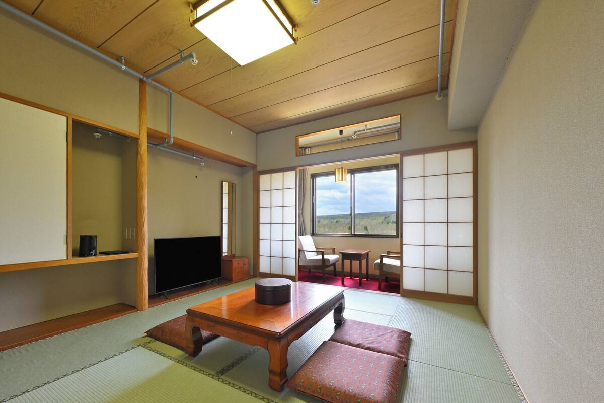Une pièce de style japonais traditionnel, avec un sol recouvert de tatamis, un téléviseur, une petite table basse et une vue sur l’extérieur.