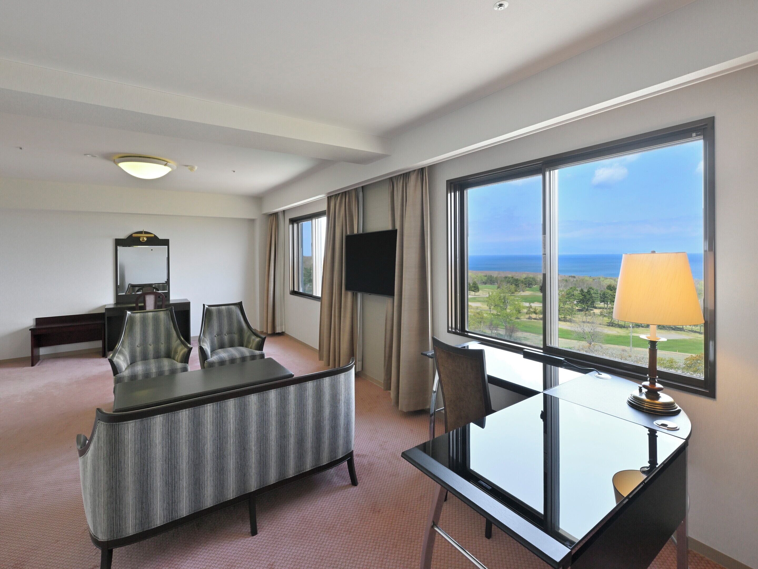 A hotel room with a bed, a desk, a chair, a TV, and a large window with a view of the ocean.