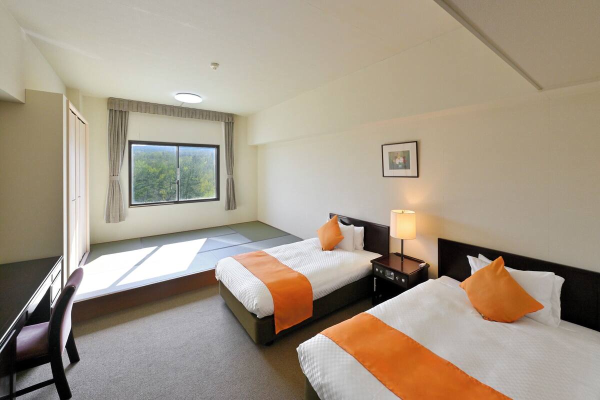 Une chambre d’hôtel avec deux lits, un bureau, une chaise, une table de chevet avec une lampe, une fenêtre avec des rideaux et un tableau encadré au mur.