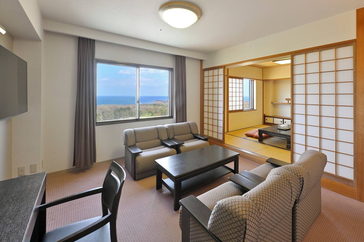 Une chambre d’hôtel équipée d’un canapé, d’une table à manger, d’une télévision, d’une grande fenêtre offrant une vue et d’une porte coulissante donnant également sur la vue.