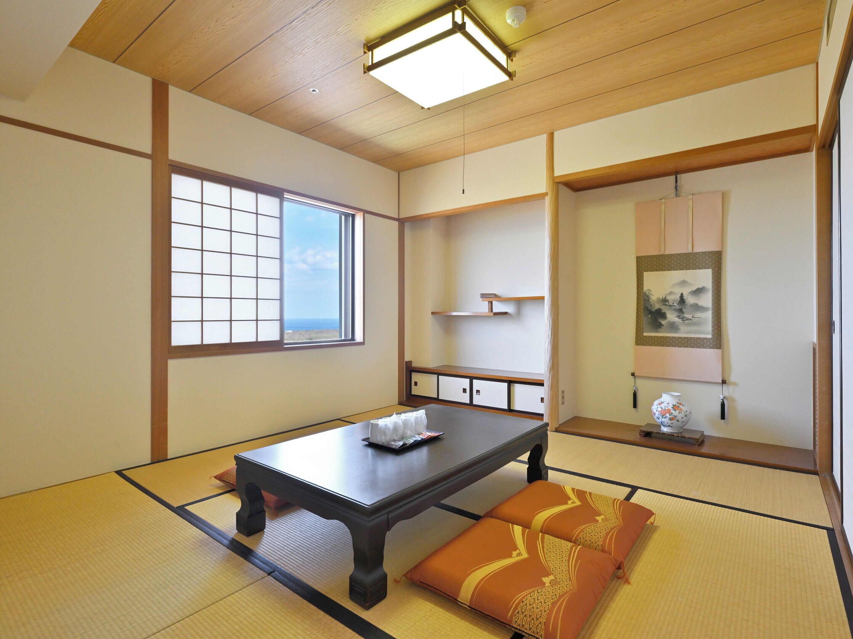 Um quarto japonês tradicional com tatames, uma mesa baixa, almofadas, uma janela com vista e uma pintura na parede.