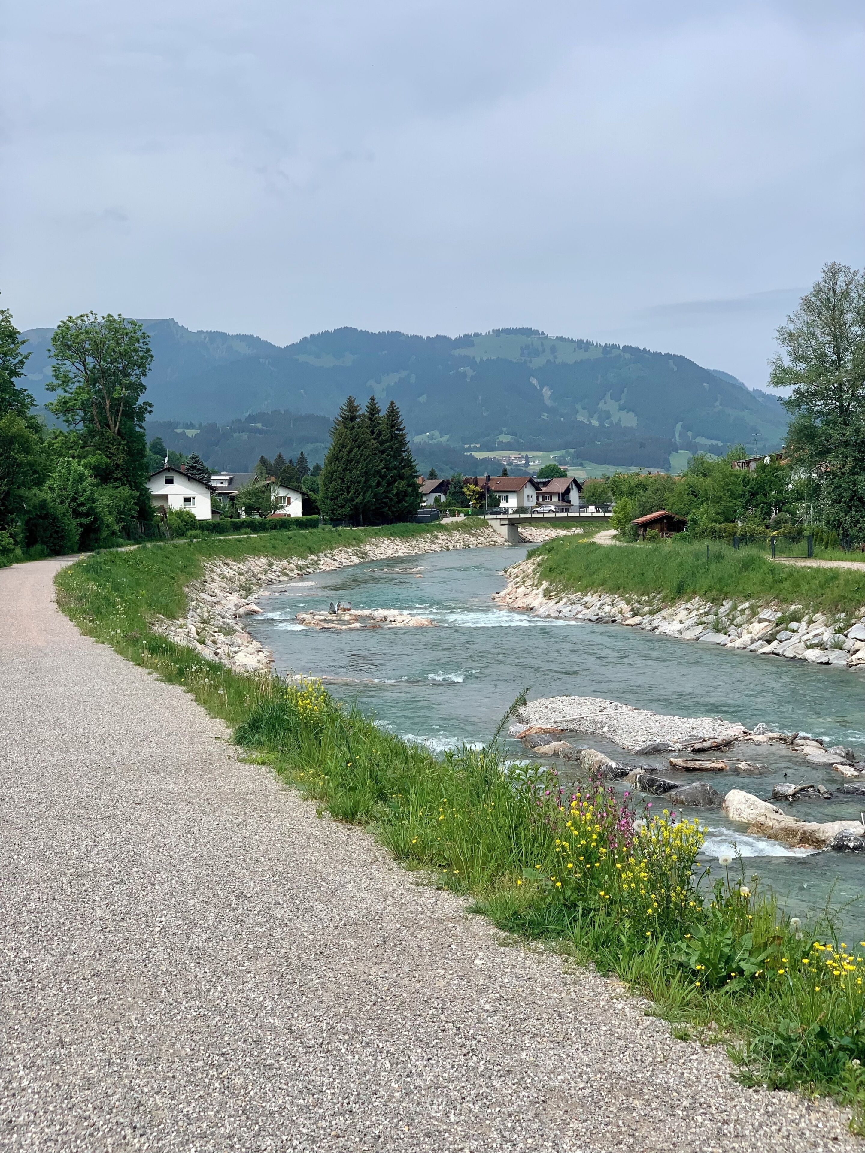 A gravel road alongside a river with houses and mountains in the background.