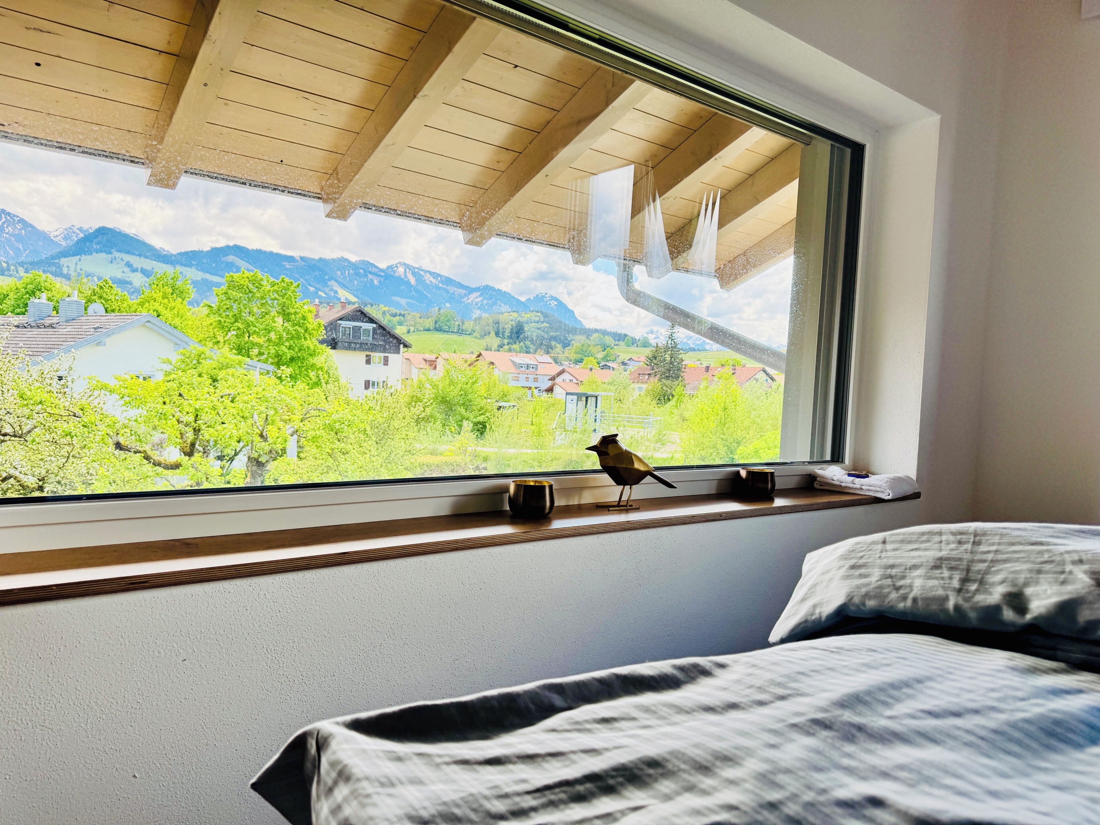 A room with a bed, a window with a view of mountains and houses, and a bird on the windowsill.