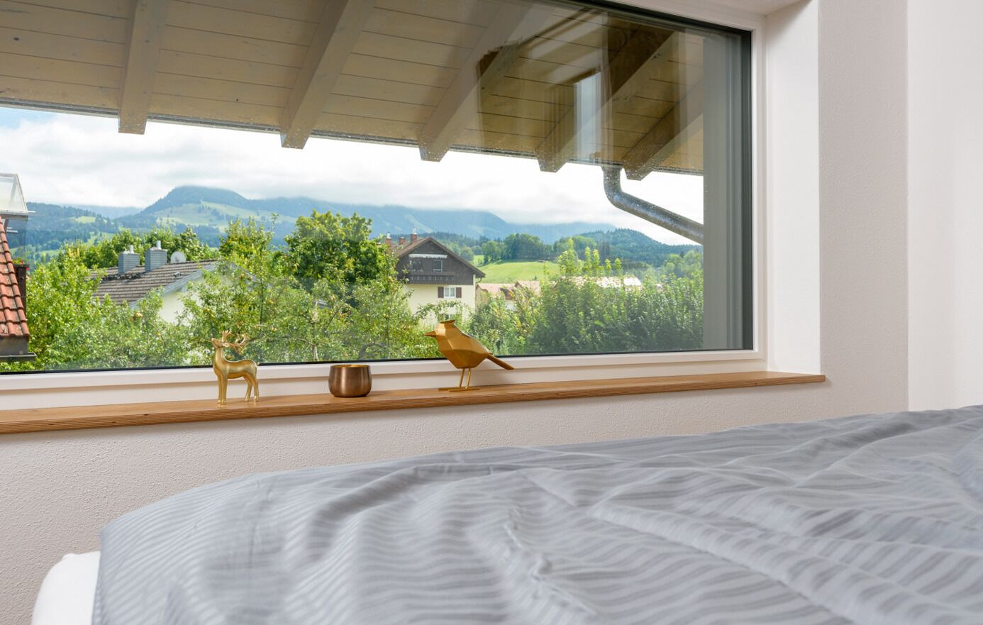 A bedroom with a large window, a bed, a wooden shelf with a deer figurine and a bird figurine, and a view of a mountain landscape.