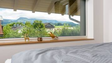 A bedroom with a large window, a bed, a wooden shelf with a deer figurine and a bird figurine, and a view of a mountain landscape.