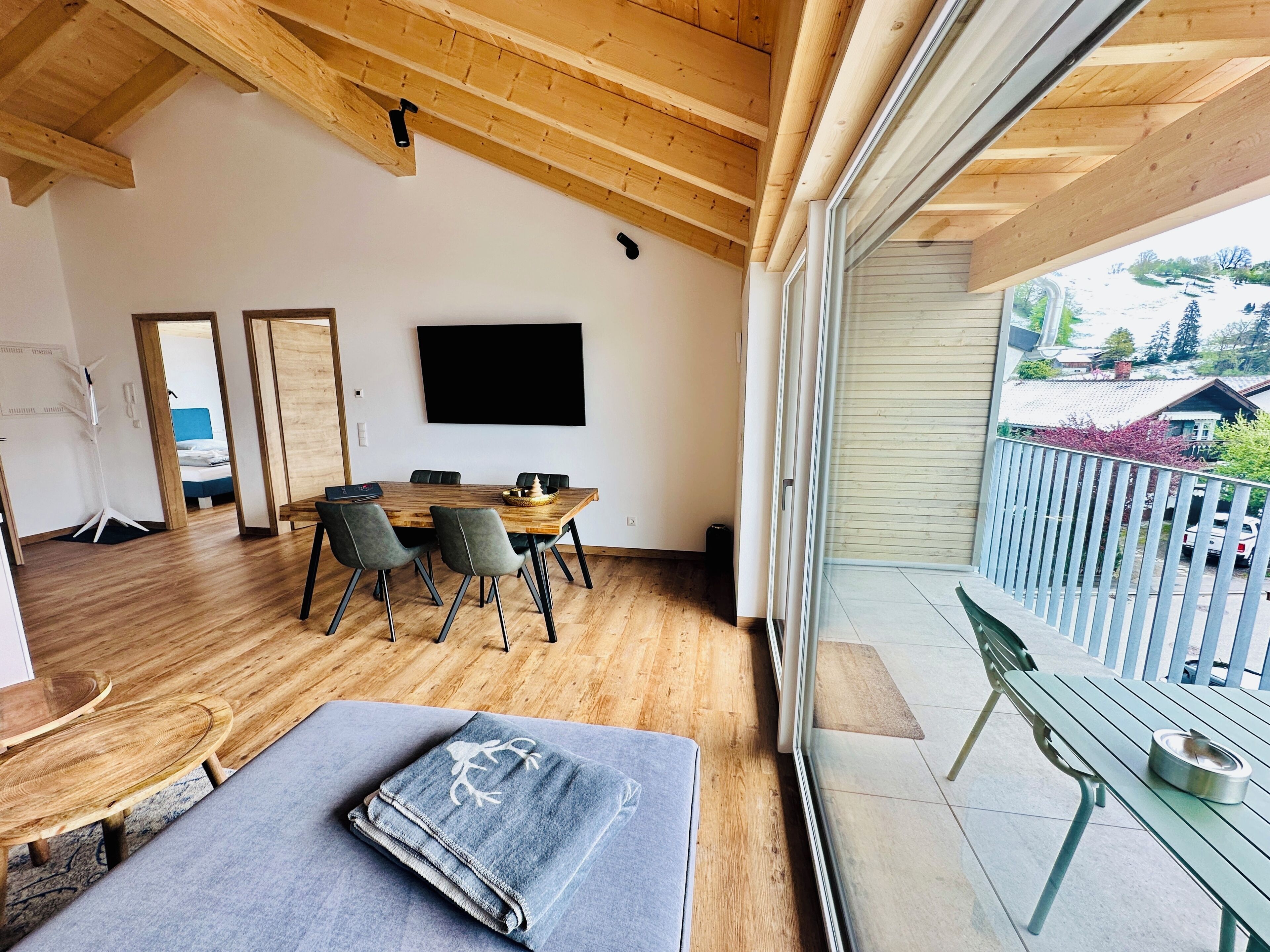 A modern interior with a wooden ceiling, a dining area with a table and chairs, a flat-screen TV mounted on the wall, and a balcony with a railing.