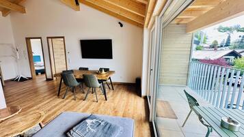 A modern interior with a wooden ceiling, a dining area with a table and chairs, a flat-screen TV mounted on the wall, and a balcony with a railing.