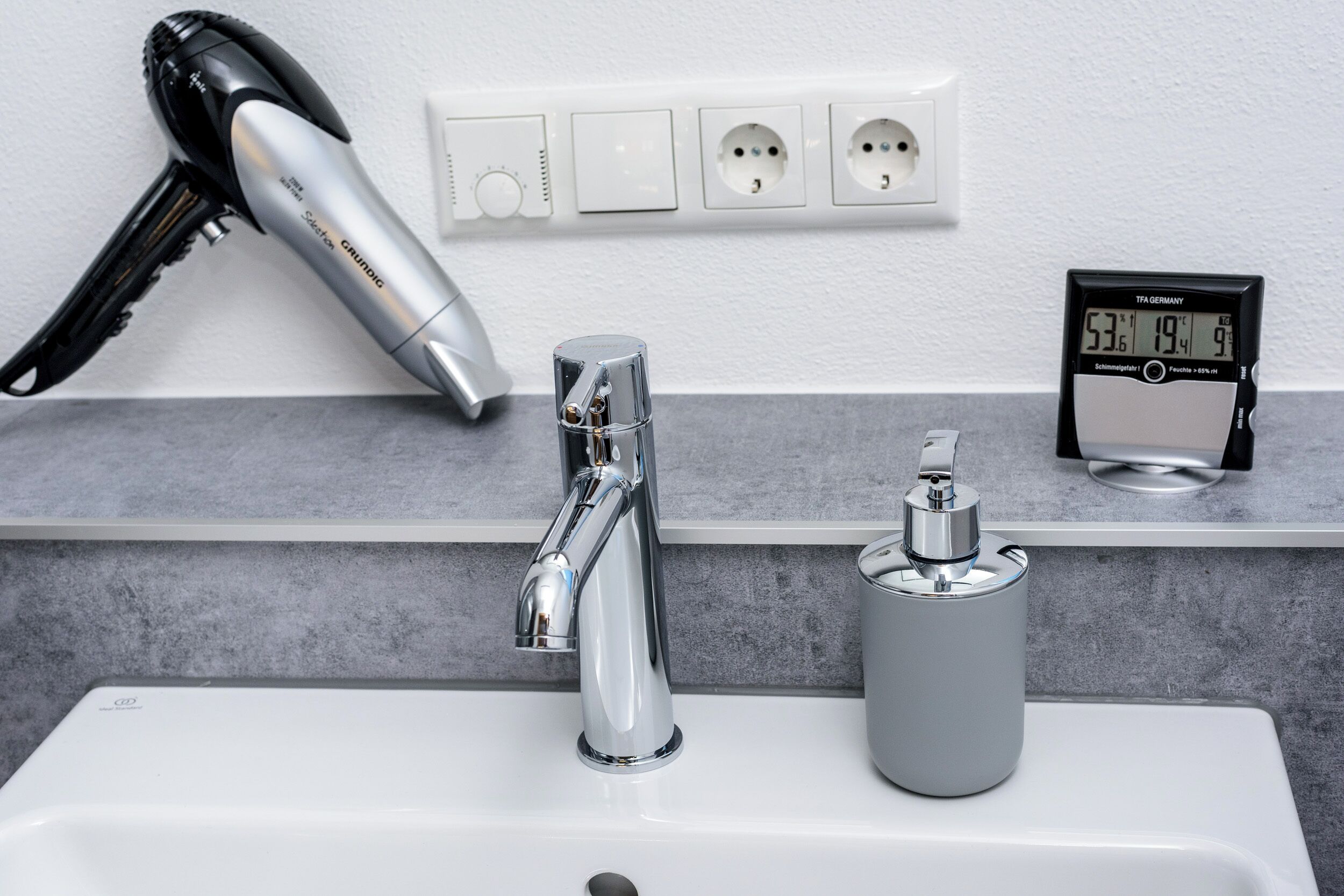 Un baño con un lavamanos moderno, un secador de pelo, un termómetro digital y un enchufe.