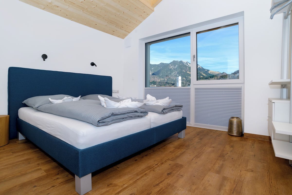 Une chambre à coucher avec un lit, une fenêtre donnant sur les montagnes, un plancher en bois et une commode blanche.