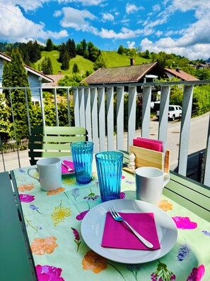 A balcony with a table set for two, featuring a floral tablecloth, two blue glass cups, a white mug, and a plate with a fork and napkin. - Berg Fux Ferienhaus und Wohnungen (Sonthofen)