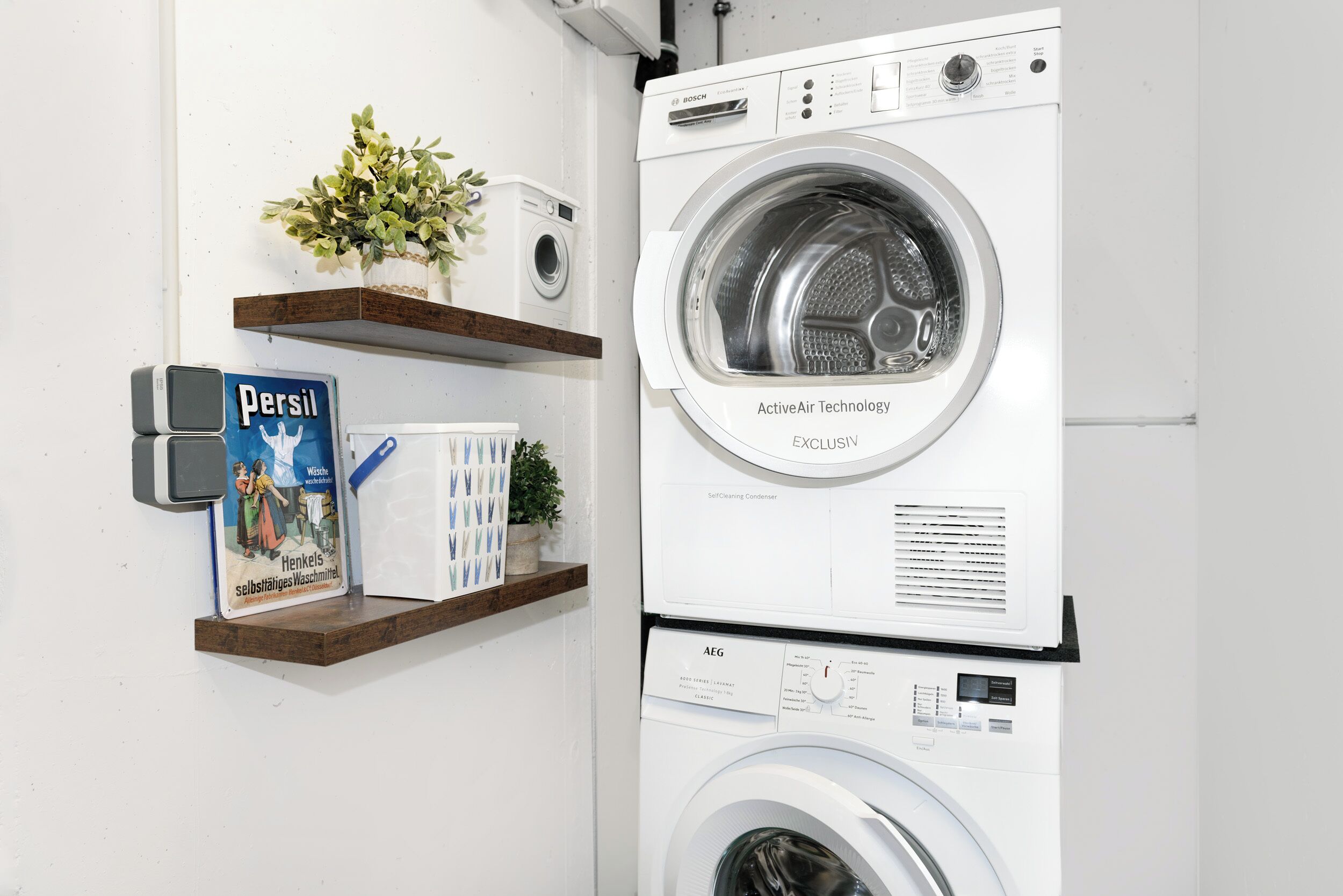 Una lavadora y secadora apiladas con un panel de control, una caja de detergente Persil y plantas decorativas en un estante.