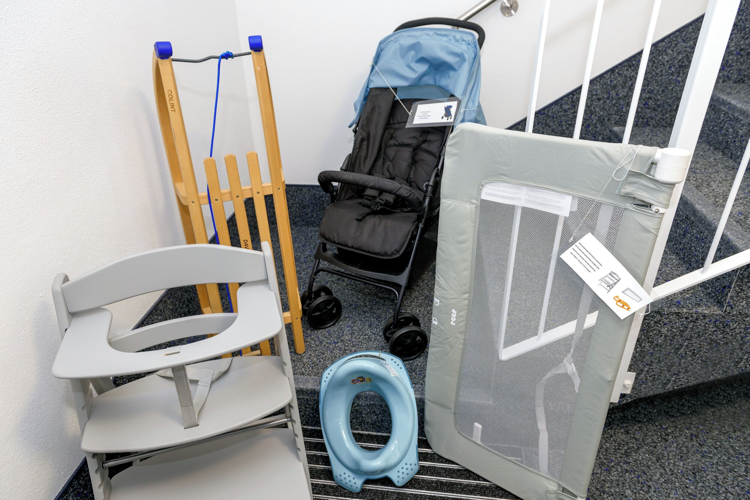 Ein Zimmer mit einem weißen Stuhl, einem blauen Toilettendeckel, einem Kinderwagen und einem Kinderzaun.