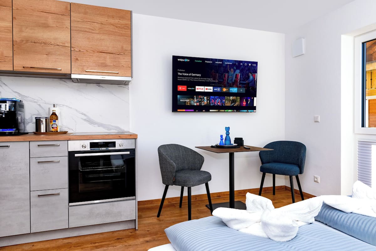 Une cuisine moderne avec des armoires en bois, un four encastré et un téléviseur à écran plat diffusant un service de streaming. On y trouve un petit coin repas avec une table et deux chaises.