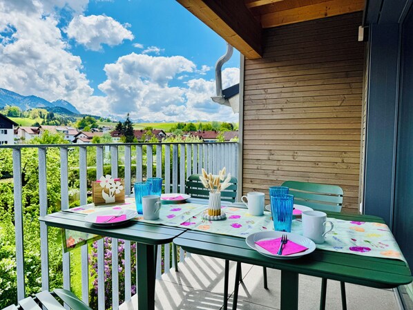 A balcony with a table set for two, a wooden wall, and a view of houses and mountains. - Berg Fux Ferienhaus und Wohnungen (Sonthofen)
