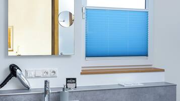 A bathroom with a sink, mirror, hairdryer, and window with blinds.