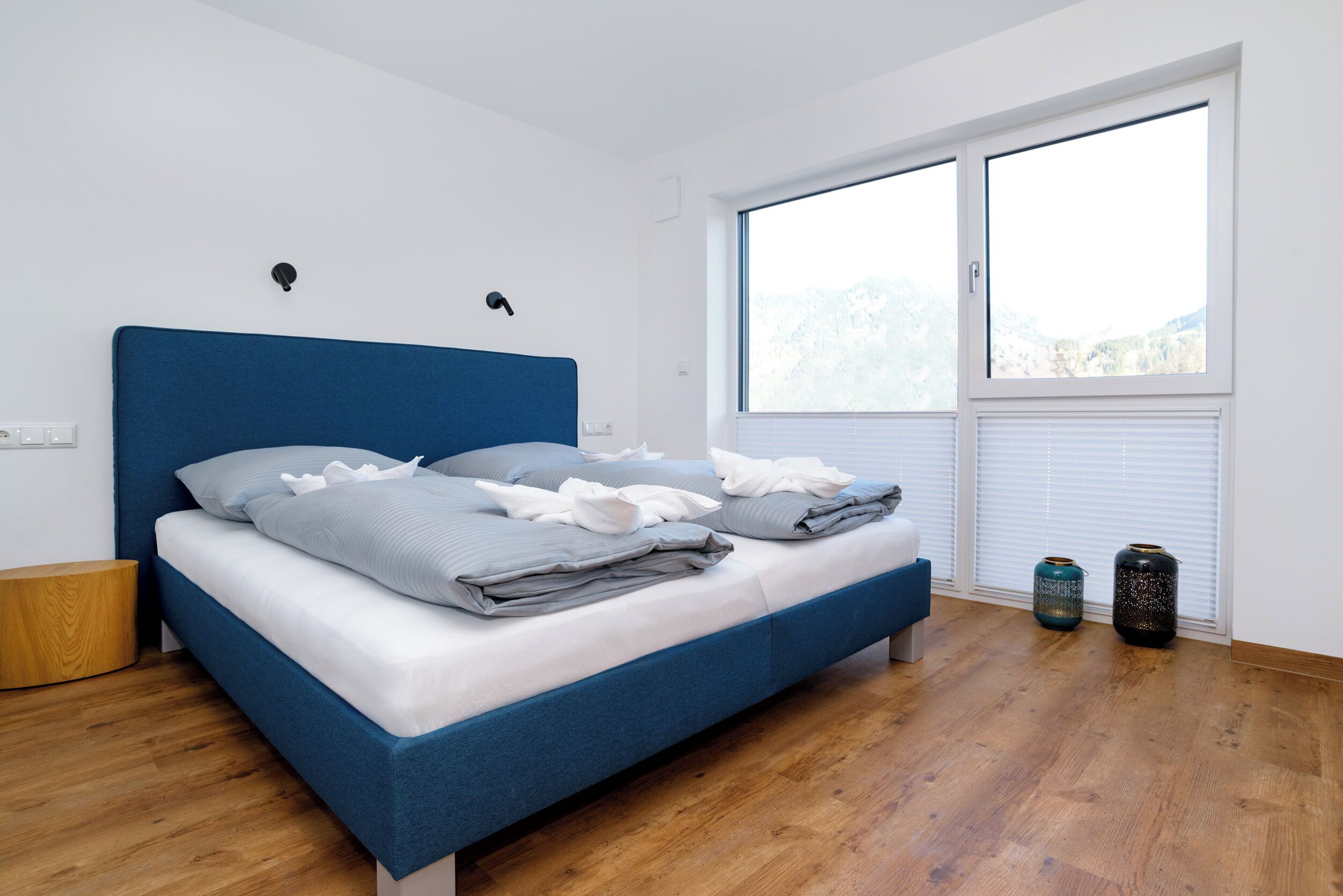 A bedroom with a blue bed, white walls, a wooden floor, and a large window with a view.