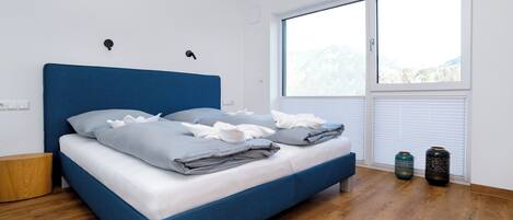 Een slaapkamer met een blauw bed, witte wanden, een houten vloer en een groot raam met uitzicht.