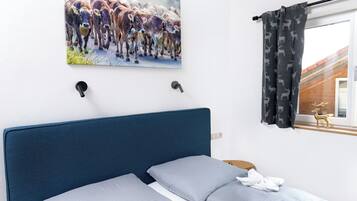 Ein Schlafzimmer mit einem Bett, einem Kuhbild, einem Fenster mit Vorhängen und Blick auf ein Gebäude.