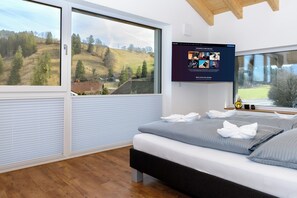 A modern bedroom with a large bed, a TV mounted on the wall, and a scenic view through the windows. - Berg Fux Ferienhaus und Wohnungen (Sonthofen)