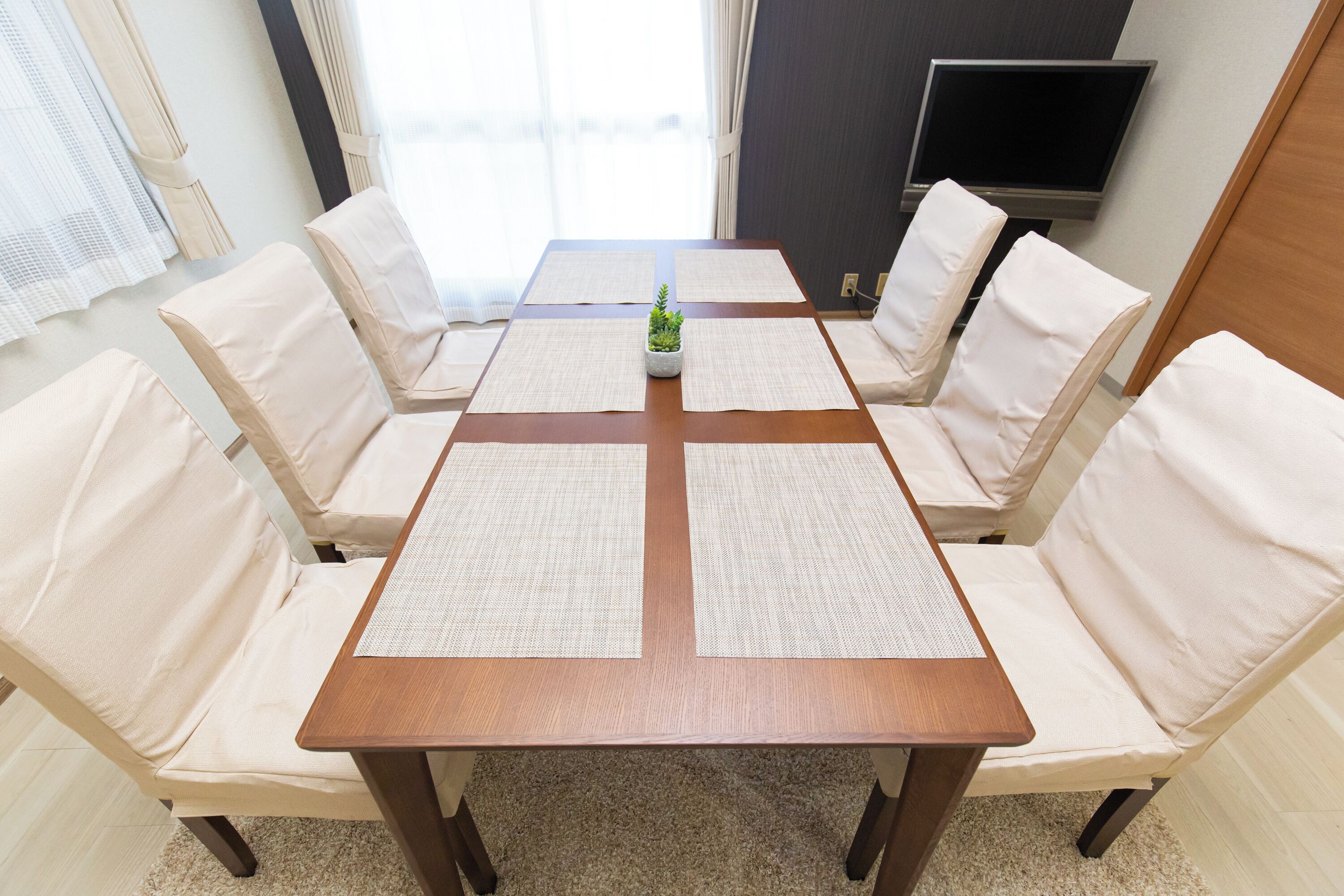 A dining area with a wooden table and six beige chairs, a small plant on the table, and a TV in the background.