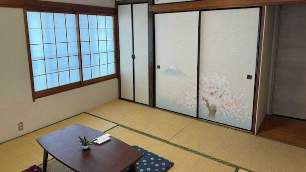 Ein traditioneller japanischer Raum mit Tatami-Bodenbelag, einer Schiebetür mit Kirschblütenmuster, einem fensterartigen Gitter und einem kleinen Tisch mit Pflanze und Buch.