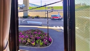 Blick aus dem Fenster auf eine Straße mit Brücke und einem Turm in der Ferne, ein Blumenbeet mit violetten Blumen und ein rotes Fahrzeug.
