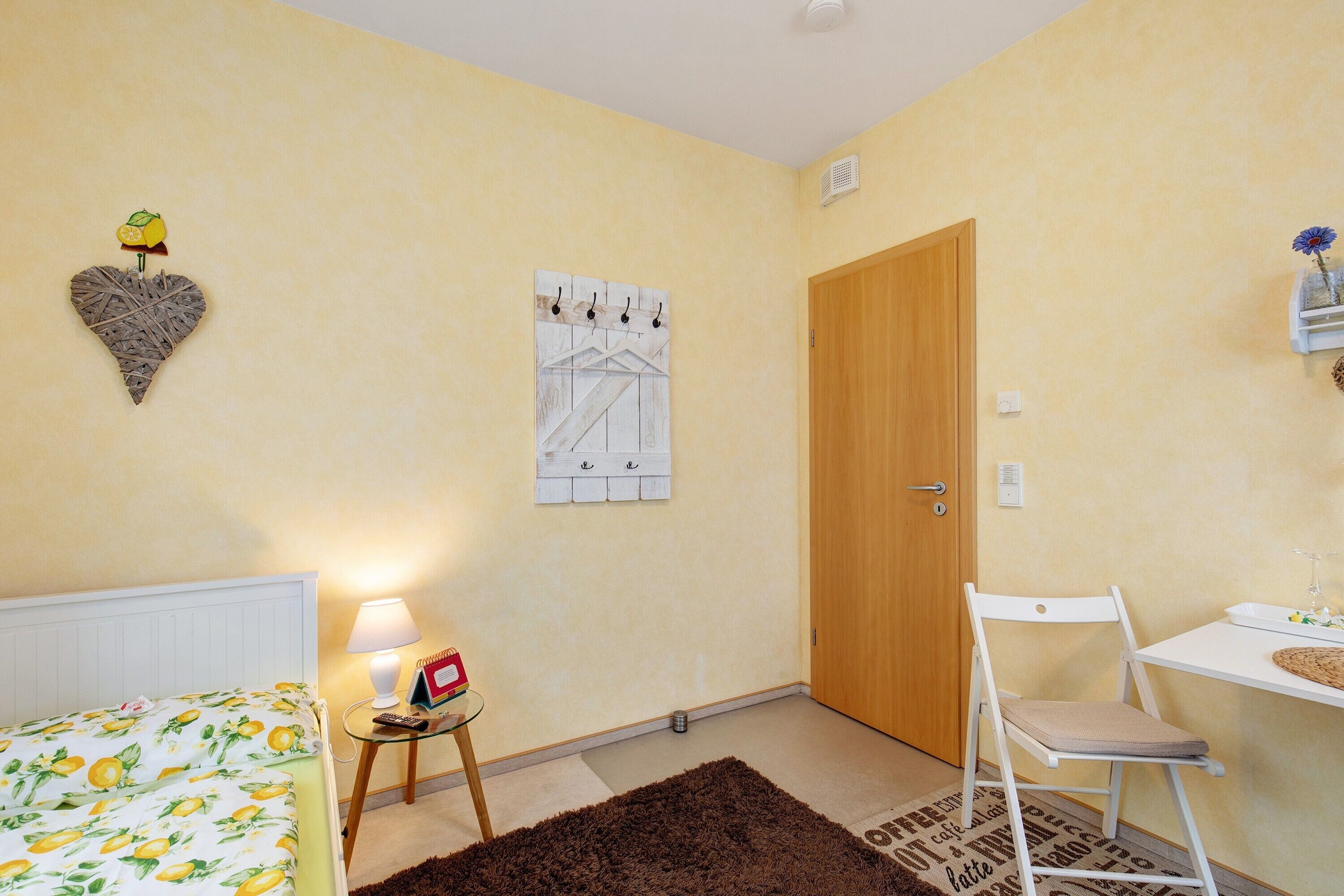 Ein Zimmer mit einem Bett, einem kleinen Tisch mit Lampe, einem Sessel, einer Tür und einem an der Wand hängenden Kalender.